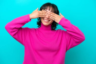 Arjantinli genç bir kadın mavi arka planda izole edilmiş. Gözlerini elleriyle kapatıyor ve gülümsüyor.