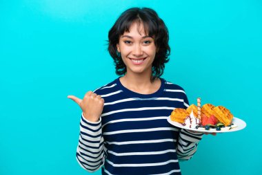 Young Argentinian woman holding waffles isolated on blue background pointing to the side to present a product