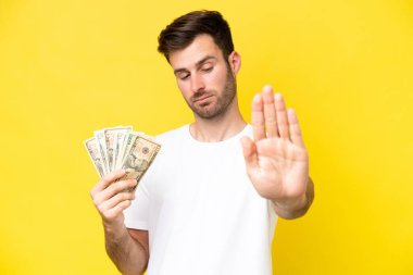 Kafkasyalı genç adam sarı arka planda izole edilmiş bir sürü para alıyor. Durma hareketi yapıyor ve hayal kırıklığına uğruyor.