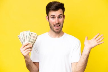 Kafkasyalı genç adam, sarı arka planda izole edilmiş ve şok olmuş bir yüz ifadesiyle bir sürü para alıyor.