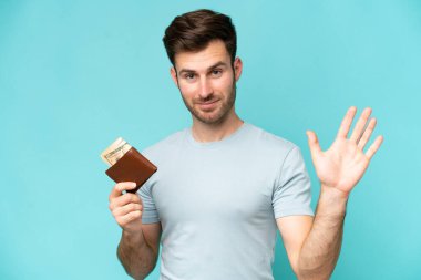 Mavi arka planda cüzdan tutan genç beyaz adam mutlu bir ifadeyle selamlıyor.