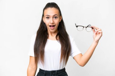 İzole edilmiş beyaz arka plan üzerinde gözlük tutan genç Arap kadın şaşırmış ve şok olmuş bir yüz ifadesi var.