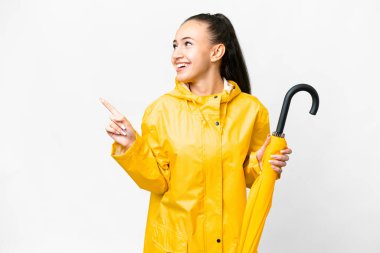 Yağmura dayanıklı ceketi ve şemsiyesi olan genç bir Arap kadın izole edilmiş beyaz arka planda parmağını göstererek bir ürün sunuyor.