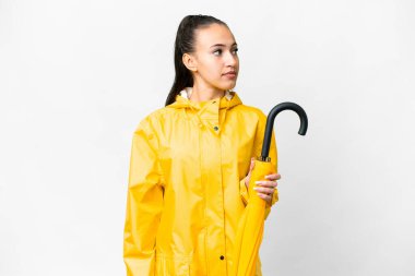 Yağmura dayanıklı paltosu ve şemsiyesi olan genç bir Arap kadın, yalıtılmış beyaz arka planda yan tarafa bakıyor.