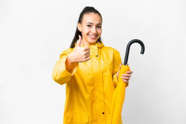 Yağmura dayanıklı ceketi ve şemsiyesi olan genç bir Arap kadın izole edilmiş beyaz arka planda başparmakları havada çünkü iyi bir şey oldu.