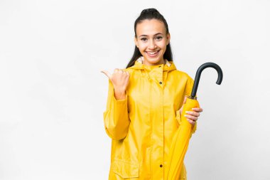 Yağmura dayanıklı ceketi ve şemsiyesi olan genç bir Arap kadını, izole edilmiş beyaz arka planda bir ürünü sunmak için kenarı işaret ediyor.