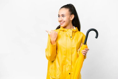Yağmura dayanıklı ceketi ve şemsiyesi olan genç bir Arap kadını, izole edilmiş beyaz arka planda bir ürünü sunmak için kenarı işaret ediyor.