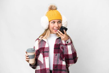 Young pretty blonde woman wearing winter muffs over isolated white background holding coffee to take away and a mobile