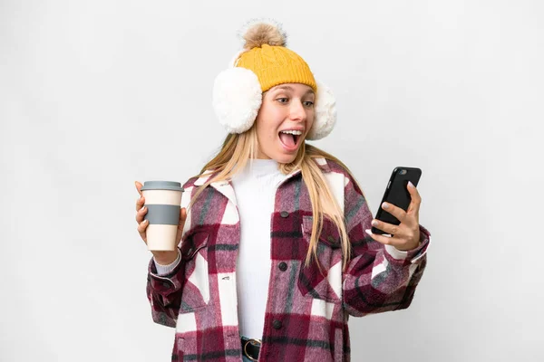 stock image Young pretty blonde woman wearing winter muffs over isolated white background holding coffee to take away and a mobile