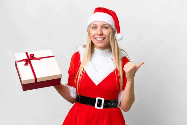年轻的金发女郎头戴圣诞礼帽 带着礼物 带着孤零零的白色背景 指着侧面展示自己的产品 — 图库照片