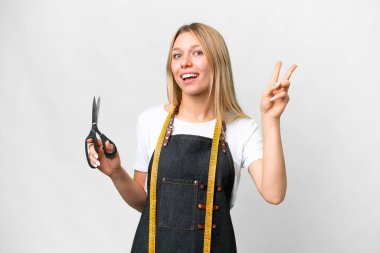 İzole edilmiş beyaz arka planda genç sarışın terzi kadın gülümsüyor ve zafer işareti gösteriyor.