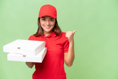 Pizza delivery caucasian woman with work uniform picking up pizza boxes isolated on green chroma background pointing to the side to present a product