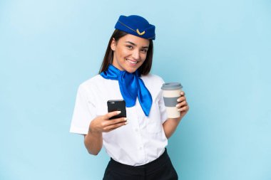 Uçak hostesi Kafkas kadın mavi arka planda izole edilmiş. Elinde kahve ve cep telefonu var.