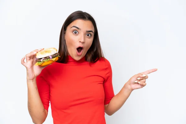 Młoda Kaukaska Kobieta Trzyma Hamburgera Izolowane Białym Tle Zaskoczony Wskazując — Zdjęcie stockowe