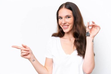 Beyaz arka planda izole edilmiş ev anahtarlarını tutan beyaz saçlı genç bir kadın şaşırmış ve parmağıyla kenarı işaret ediyor.