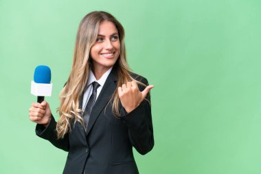 Genç ve güzel Uruguaylı televizyon sunucusu izole edilmiş bir arkaplanda bir ürünü sunmak için kenarı işaret ediyor.