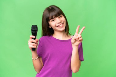 Küçük şarkıcı kız izole edilmiş bir arka planda mikrofonu kaldırıyor gülümsüyor ve zafer işareti gösteriyor