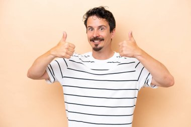 Beyaz tenli genç adam bej arka planda izole edilmiş bir şekilde baş parmak işareti yapıyor.