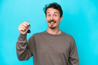 Young caucasian man holding home keys isolated on blue background with surprise and shocked facial expression