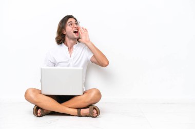 Yerde dizüstü bilgisayarı olan yakışıklı genç bir adam beyaz arka planda izole edilmiş ve ağzı sonuna kadar açık bağırıyor.