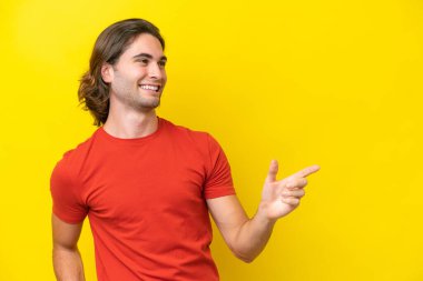 Beyaz tenli yakışıklı adam sarı arka planda izole edilmiş. Parmakla işaret ediyor ve bir ürün sunuyor.