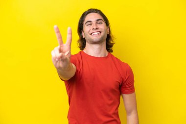 Sarı arka planda izole edilmiş beyaz yakışıklı adam gülümsüyor ve zafer işareti gösteriyor.