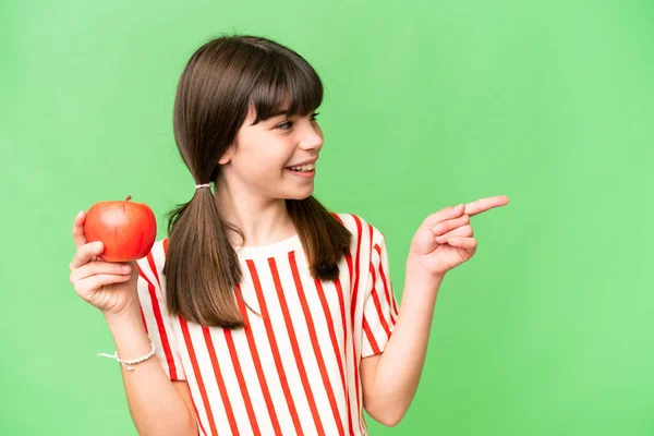 Little caucasian girl holding an apple over isolated background pointing to the side to present a product
