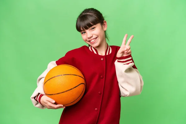 在孤独的背景下打篮球的高加索小女孩微笑着 并展示出胜利的迹象 — 图库照片
