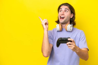 Sarı arka planda izole edilmiş bir bilgisayar oyunu kontrolörüyle oynayan yakışıklı beyaz bir adam çözümü bulmak için parmağını kaldırıyor.