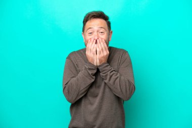 Mavi arka planda izole edilmiş orta yaşlı beyaz bir adam mutlu ve gülümseyen elleriyle ağzını kapatıyor.