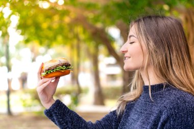 Genç ve güzel Romen kadın elinde neşeli bir ifadeyle açık havada hamburger tutuyor