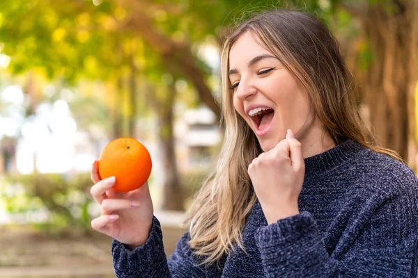 stock image Young pretty Romanian woman holding an orange at outdoors celebrating a victory