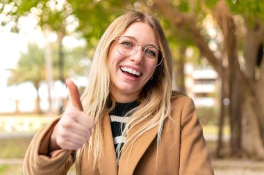Açık havada genç, güzel, sarışın bir kadın. Başparmakları havada. Çünkü iyi bir şey oldu.