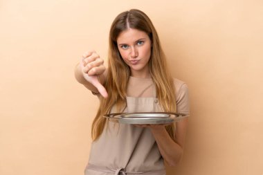 Bej arka planda izole edilmiş tepsisi olan genç sarışın garson kadın olumsuz bir ifadeyle baş parmağını aşağı gösteriyor.