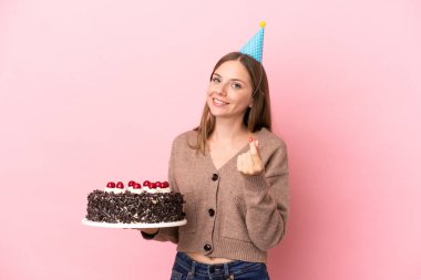 Young Lithuanian woman holding birthday cake isolated on pink background making money gesture