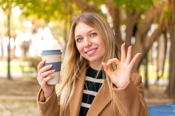 年轻漂亮的金发女人在户外拿着一杯咖啡 手举着手示意 — 图库照片
