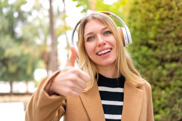 Νεαρή Όμορφη Ξανθιά Γυναίκα Ακούει Μουσική Ακουστικά Εξωτερικούς Χώρους Αντίχειρες — Φωτογραφία Αρχείου