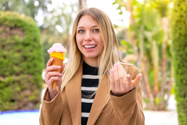若いかなりブロンドの女性が屋外でコーネットアイスクリームを持っていて 手で来るように誘います 来てくれて嬉しい — ストック写真