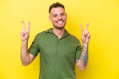 Sarı arka planda izole edilmiş beyaz, yakışıklı genç bir adam iki eliyle zafer işareti gösteriyor.