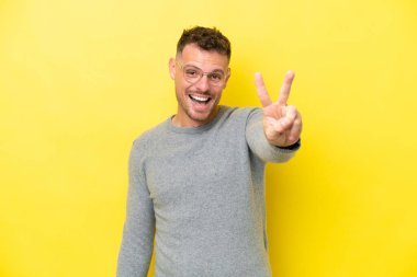 Sarı arka planda izole edilmiş yakışıklı genç adam gülümsüyor ve zafer işareti gösteriyor.