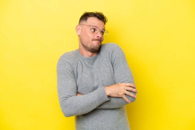 Beyaz tenli yakışıklı adam sarı arka planda izole edilmiş. Omuzlarını kaldırırken şüpheyle hareket ediyor.
