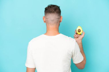 Avokado tutan beyaz bir genç arka planda mavi bir arka planda izole edilmiş.