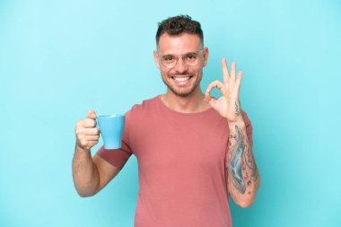 Mavi arka planda bir fincan kahve tutan genç beyaz adam parmaklarıyla tamam işareti gösteriyor.