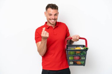 Beyaz arka planda izole edilmiş yiyecek dolu bir alışveriş sepeti tutan beyaz bir adam para hareketi yapıyor.