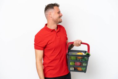 Beyaz arka planda izole edilmiş yiyecek dolu bir sepet taşıyan beyaz bir adam yan yana gülerek gülüyor.