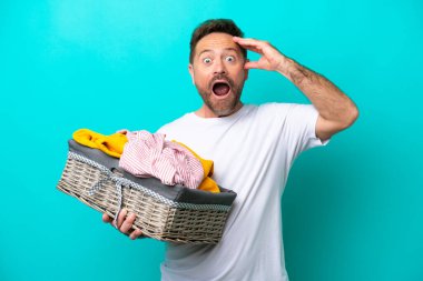 Mavi arka planda, sürpriz bir ifadeyle izole edilmiş çamaşır sepeti tutan orta yaşlı bir kadın.