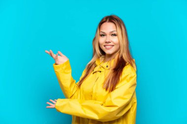 Yağmura dayanıklı bir ceket giyen genç kız izole edilmiş mavi arka planda davet edilmek için ellerini yana uzatıyor.