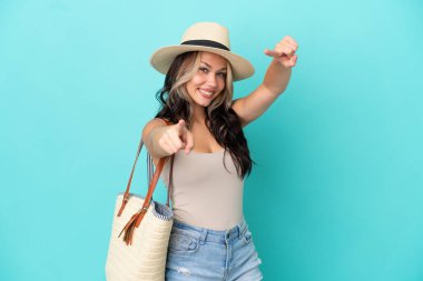 Pamel ve plaj çantalı genç Rus kız mavi arka planda izole edilmiş sana gülümserken parmak atıyor.