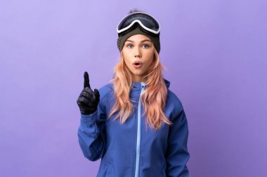İzole edilmiş mor arka planda snowboard gözlüklü genç kayakçı kız parmağını kaldırarak çözümü bulmaya çalışıyor.