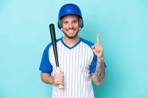 ヘルメットとバットを持つ野球選手は 最高の兆候で指を見せ 持ち上げる青い背景に隔離されています — ストック写真